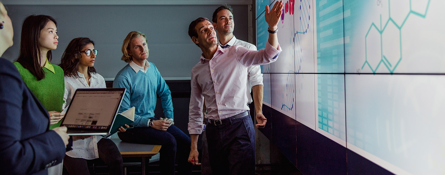 Colleagues in business casual make notes in front of a screen displaying information next to illustrations of DNA.