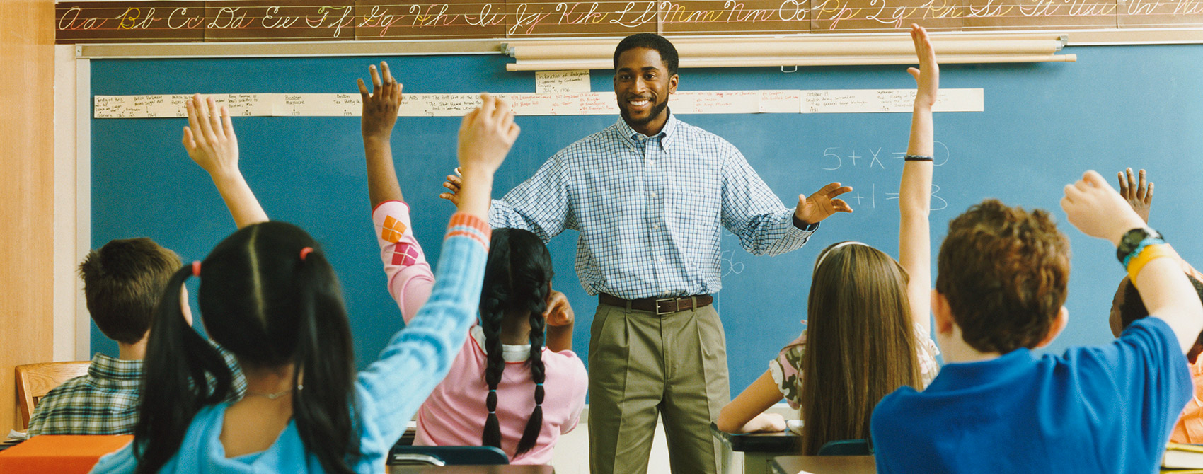 Math teacher stands in front of chalkboard with simple equations while class of diverse middle school students raise hands to answer the question.