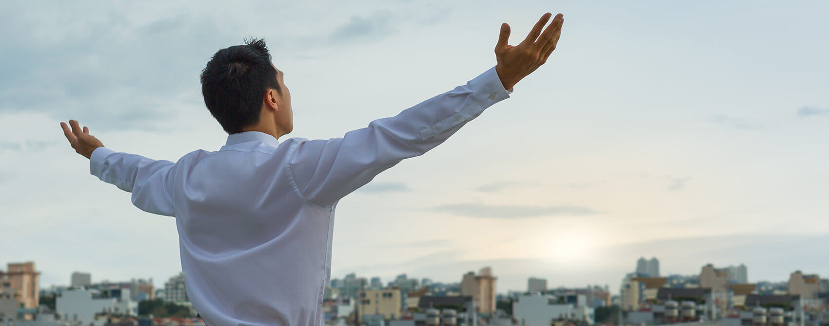 A man wearing a white button-down shirt raises his arms in supplication on a rooftop at sunrise.