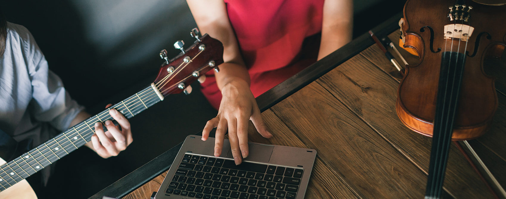 A violinist whose instrument lays on the desk works on a laptop to record her colleague playing guitar.