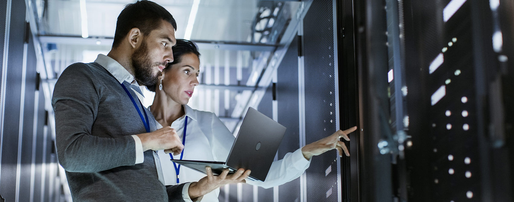 Information technology professionals examine a computer server. The male is holding a laptop, and the female is gesturing at the server.