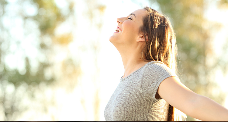 Exuberant woman smiles with her eyes closed as she raises her head toward the sky, arms out.