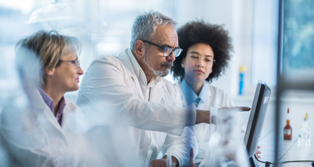 Three lab techs stand with beakers and computers utilizing forensic technology like Rapid DNA testing.