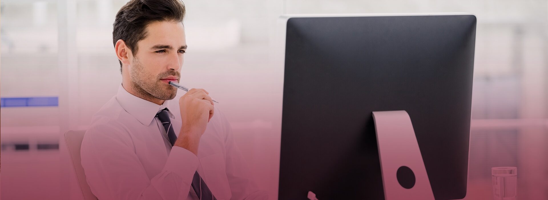 man holding a pen sitting at desk looking at computer monitor