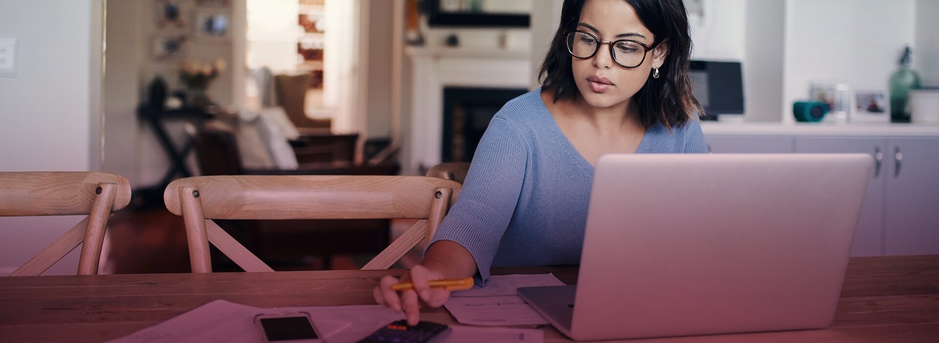 woman using calculator at dining table next to paperwork and laptop