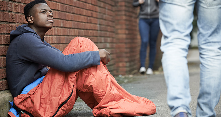 Homeless african-american teen sitting up in a sleeping bag on a busy sidewalk.