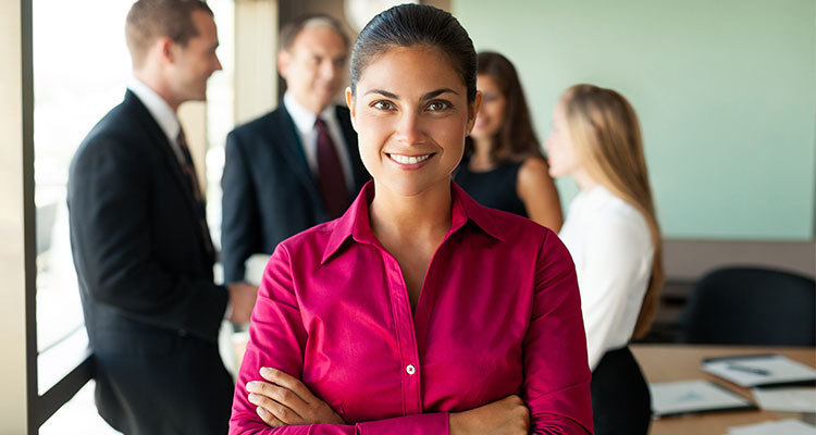 Female in business attire smiles with her arms folded while colleagues talk in the background.