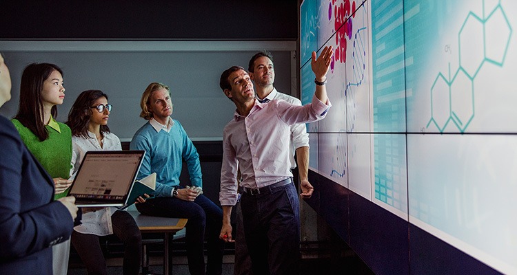 Man wearing business casual explains medical biotechnology product plans to colleagues on a large wall screen.