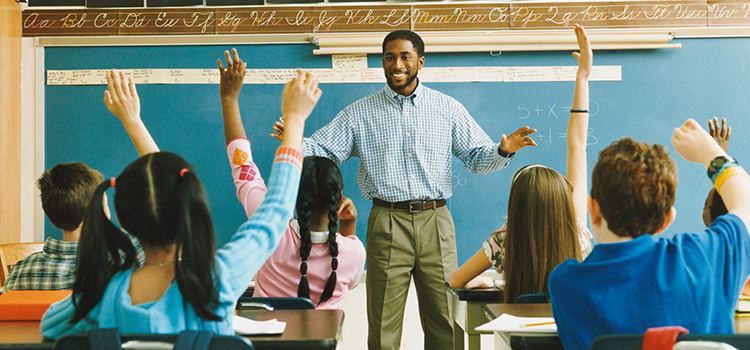 Photo of a teacher smiling at the front of an elementary classroom full of students with hands raised.