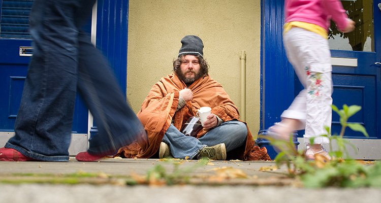 Man with unkempt hair and beard sitting on sidewalk in a blanket and jeans begging for money.